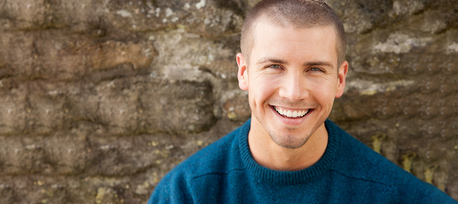 The image shows a young man with short hair, wearing a dark blue sweater, smiling and looking directly at the camera.