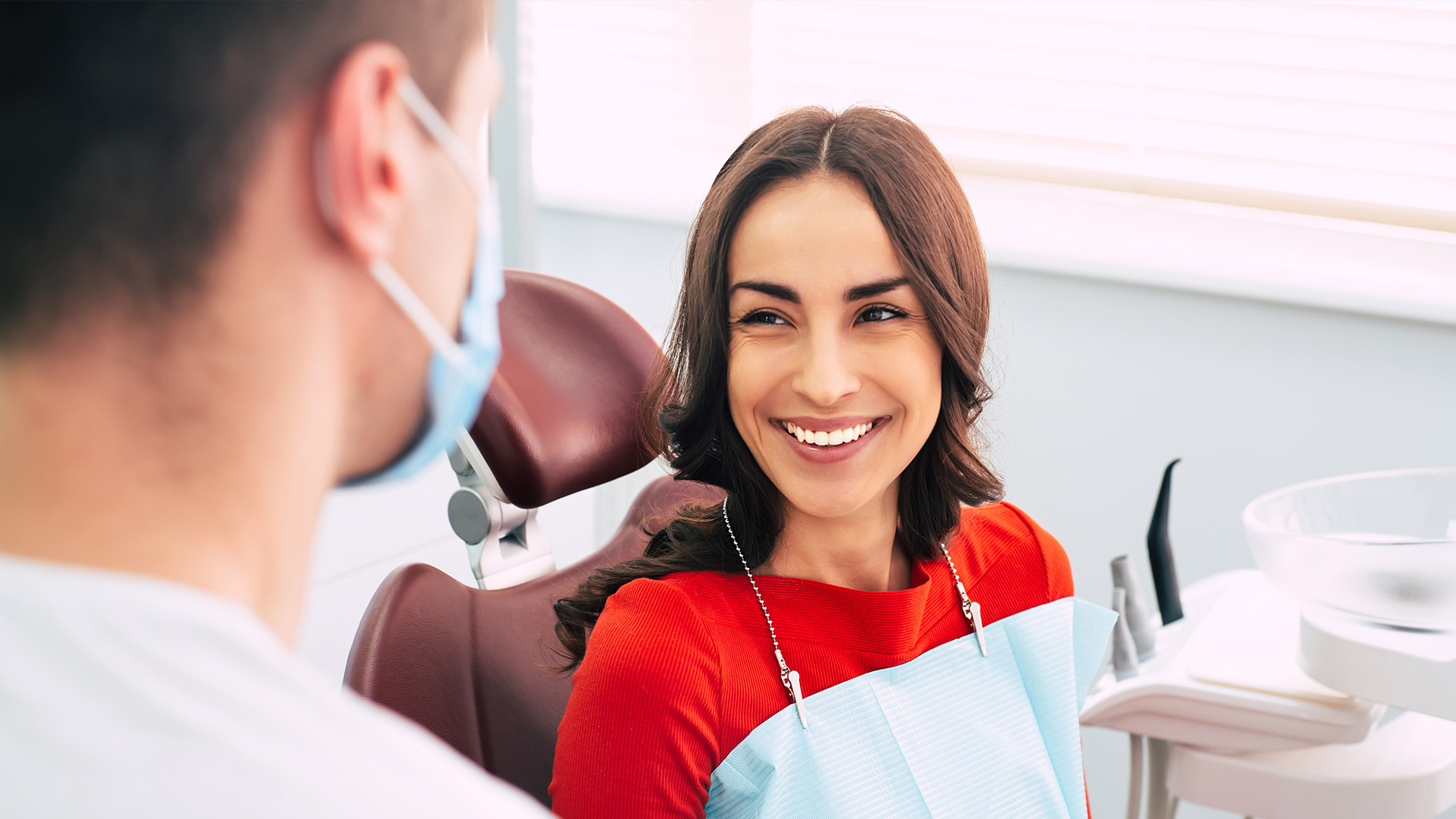 A dental professional in a white coat and mask is smiling at the camera, with a patient seated in a chair behind her.