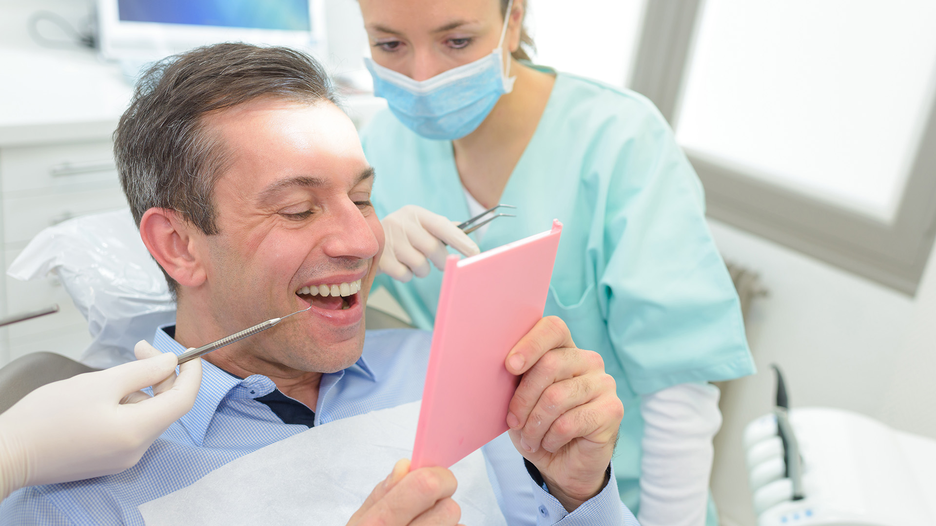 A man in a dental chair, holding a pink card or paper, smiling, with a woman standing behind him wearing a surgical mask and gloves.