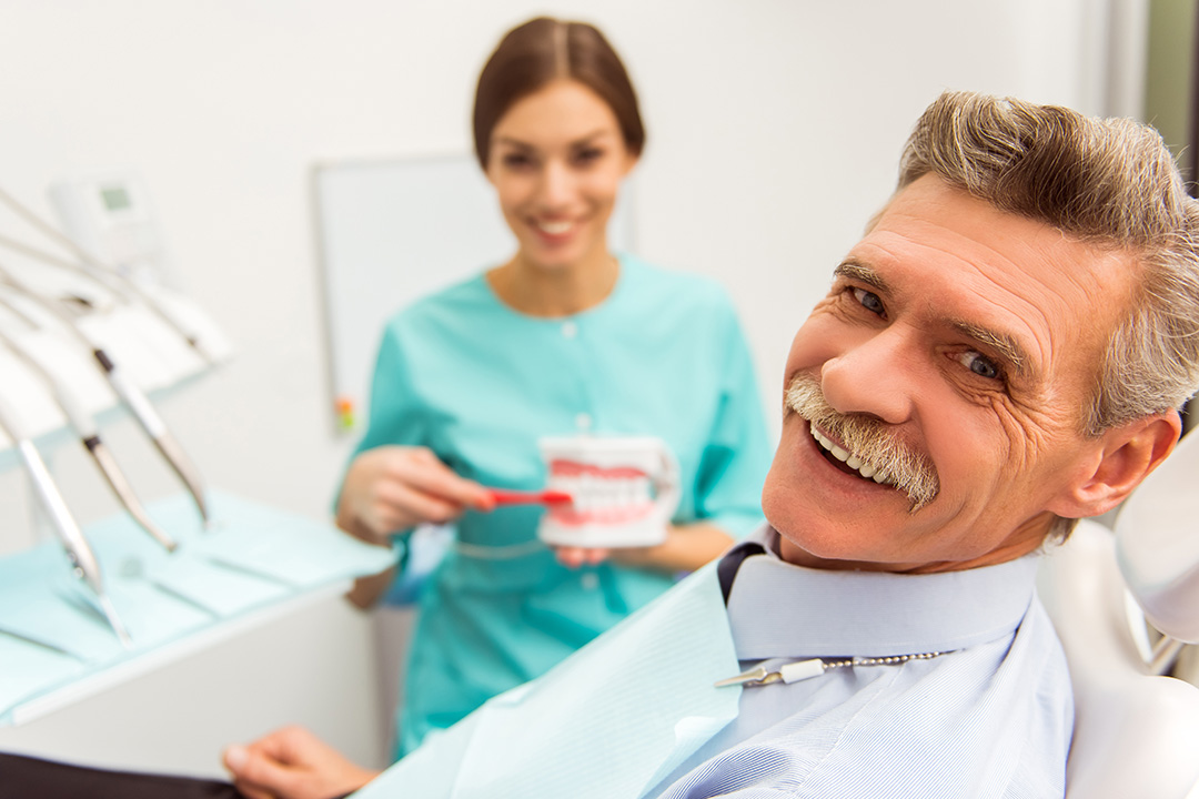 The image shows a man sitting in a dental chair with a smiling woman standing behind him, holding a toothbrush. They are both in a dental office setting.