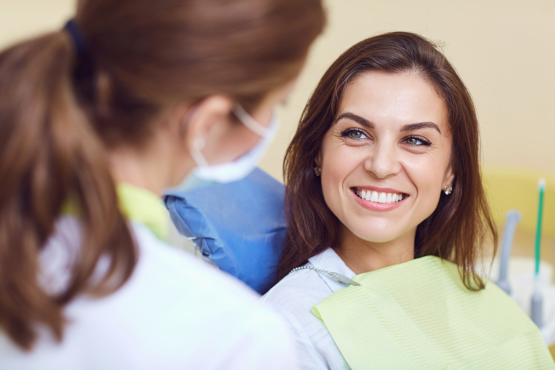A woman in a dental chair receiving dental care, smiling towards the camera, while a dentist in a white coat and mask attends to her.