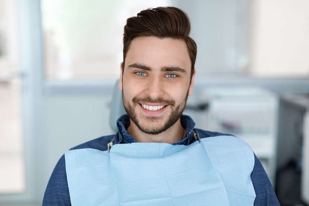 A man with a beard and blue eyes is seated in a dental chair, smiling at the camera while wearing a blue surgical mask.