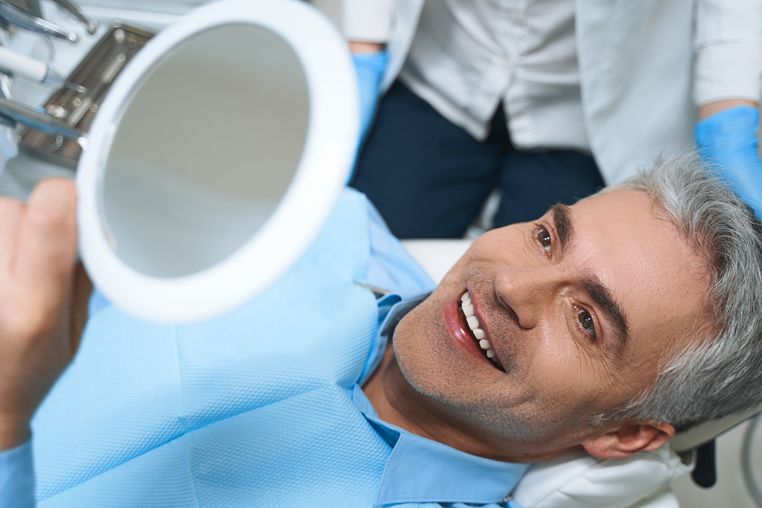 The image shows a man sitting in a dental chair, smiling at the camera, with a dental professional standing behind him, holding a mirror and examining his teeth.