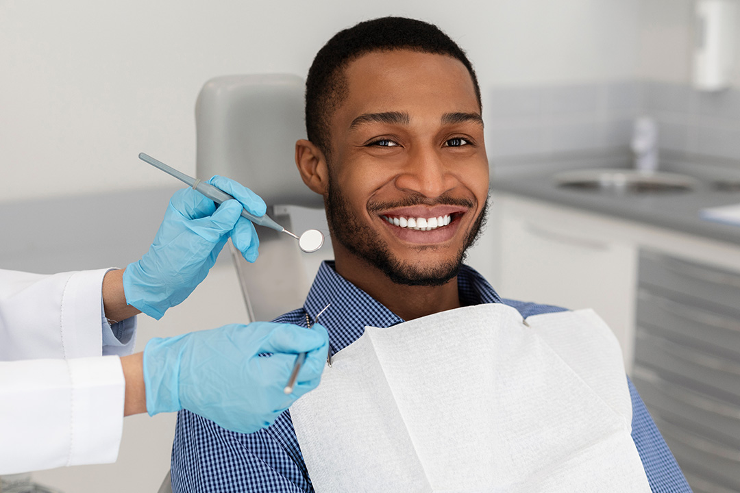 A smiling man in a dental chair receiving dental care, with a hygienist applying a dental tool to his mouth.
