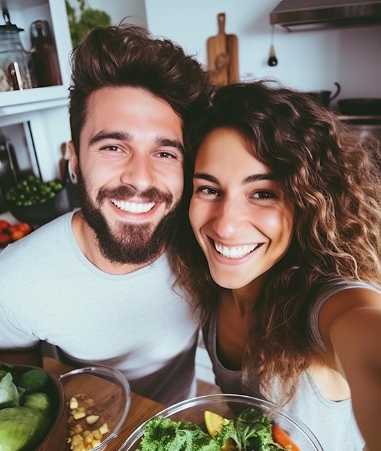 A man and a woman are smiling at the camera, standing in front of a kitchen counter with various food items.