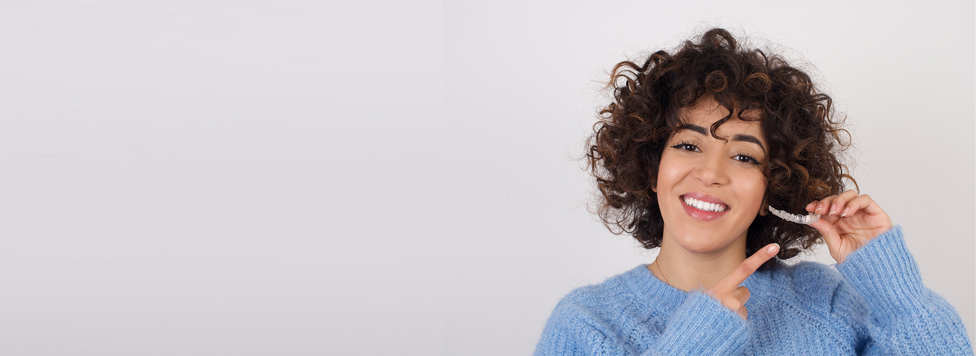 This is a color photograph of a smiling woman with curly hair, wearing a blue sweater, standing against a white background.