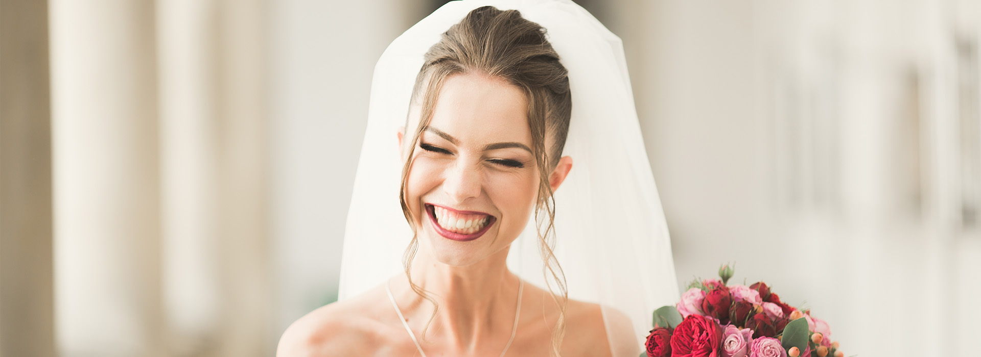 The image shows a bride in a white wedding dress, smiling and looking over her shoulder, with a veil covering her face, standing next to a bouquet of flowers.