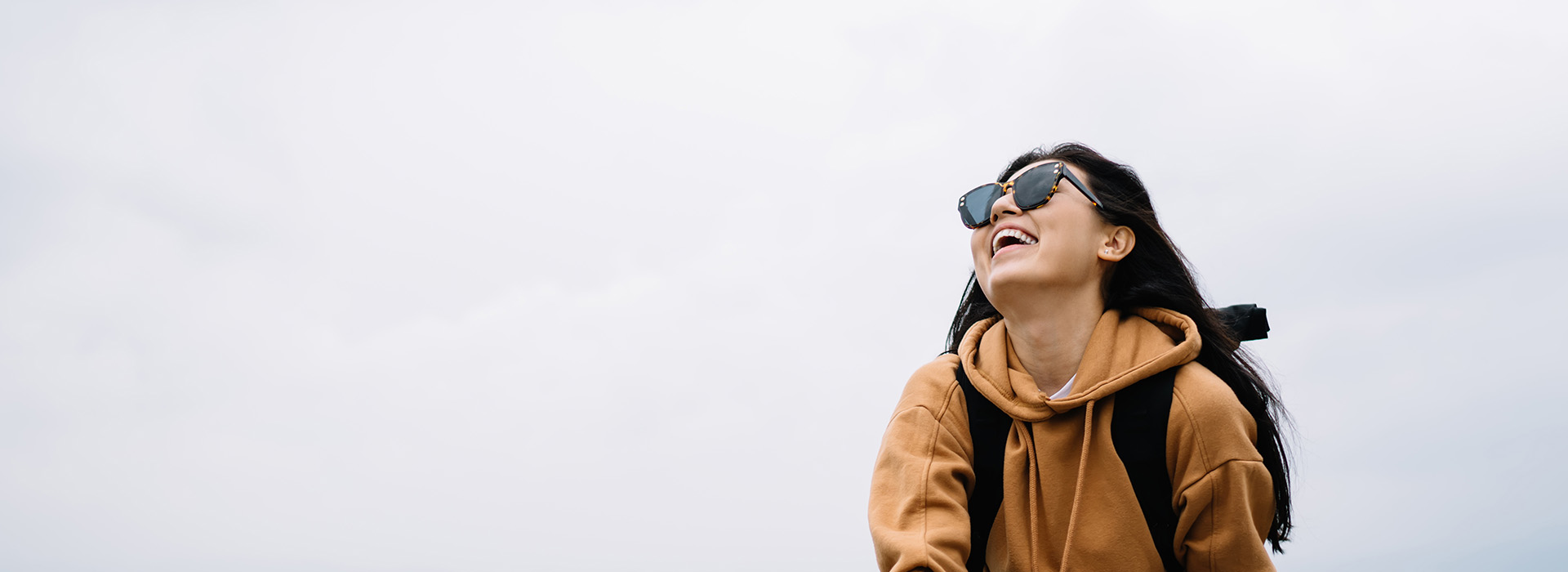 The image shows a person standing outdoors, wearing sunglasses and a backpack, with a clear sky in the background.