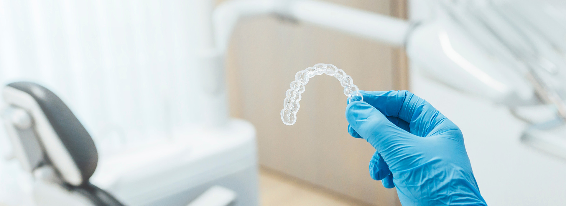 The image shows a close-up of a transparent dental implant being held by a person wearing blue gloves, with the background featuring medical equipment and a dental chair.