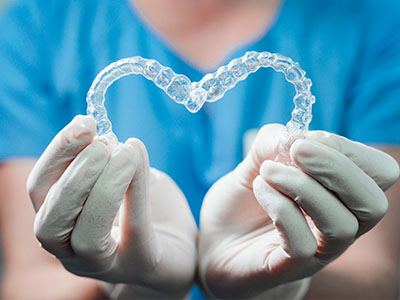 The image shows a pair of clear dental retainers held delicately in a gloved hand against a blue background.