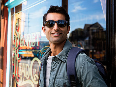 A man in sunglasses is standing outdoors, leaning against a window with his backpack on.