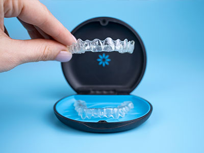 A person s hand holding a clear plastic dental retainer, showcasing its design and the blue base of the appliance, set against a light blue background.