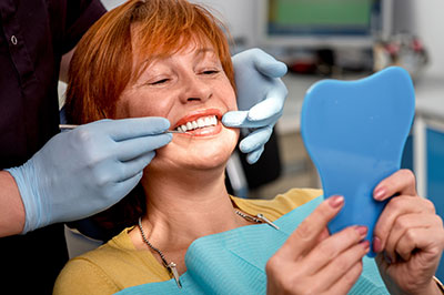 The image shows a woman seated in a dental chair, with a dental professional performing a procedure on her teeth. She is holding up a blue mouthguard and smiling at the camera.