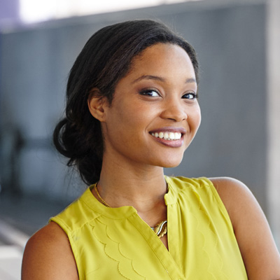 A young woman with a radiant smile, wearing a yellow top, poses confidently against a backdrop of a building.