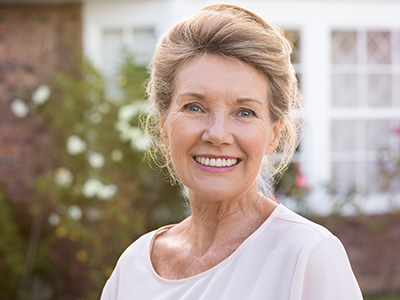 The image shows a woman standing outdoors, smiling at the camera. She has short hair and is wearing a light-colored top with a darker cardigan over it. Behind her is a house with a brick facade and a white window frame.