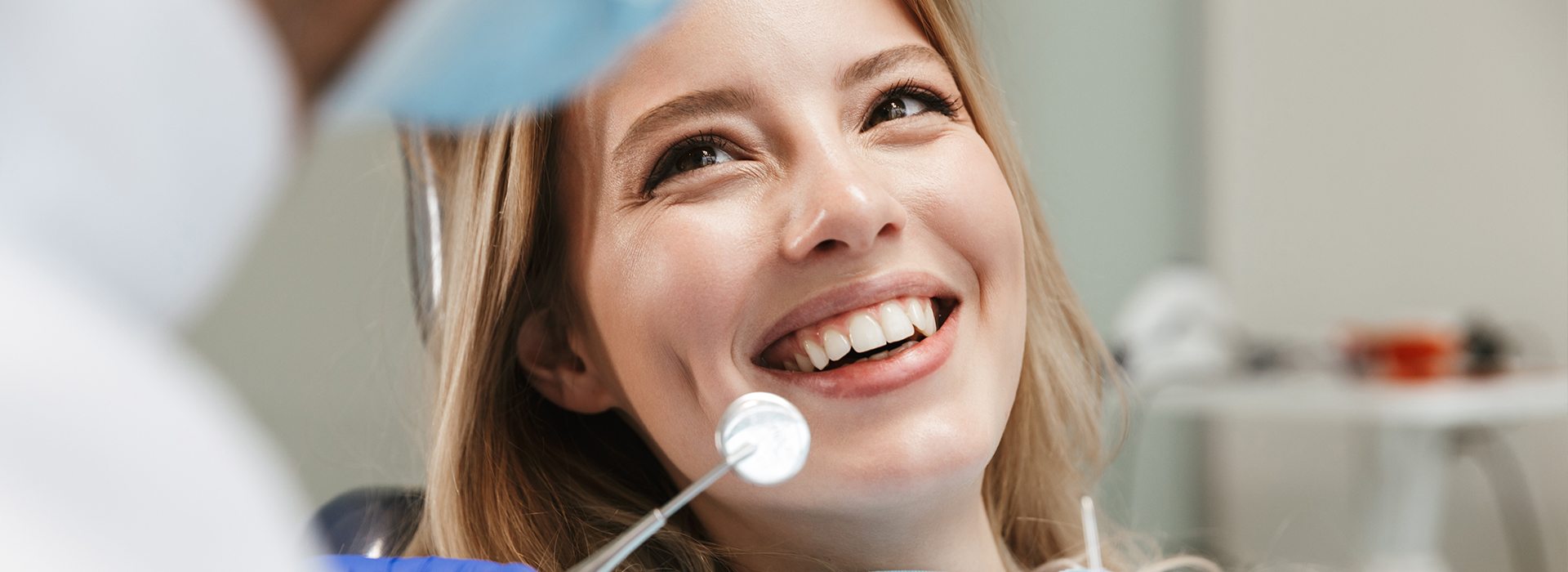 Smiling woman in dental chair, undergoing dental procedure with dental professional behind her.