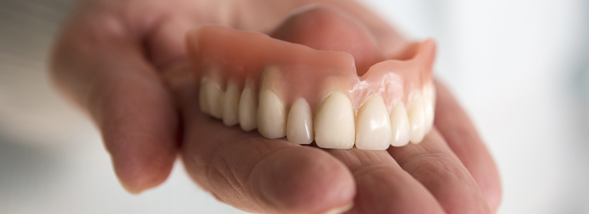 The image shows a person holding up a set of dentures against their teeth, with the focus on the artificial teeth and the contrast between them and natural teeth.