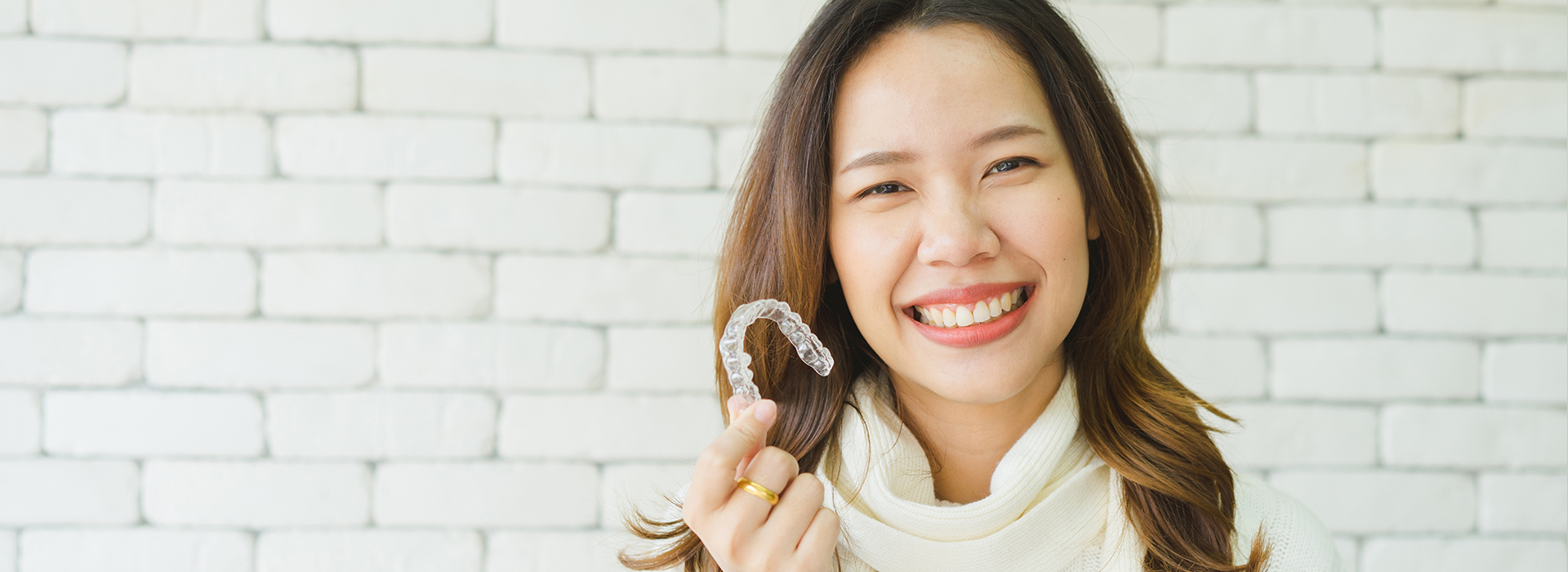 A woman with a radiant smile is holding a ring, standing against a brick wall.