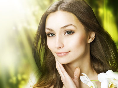 A woman with light skin, long brown hair, and a neutral expression gazes directly at the camera against a softly blurred backdrop of greenery.