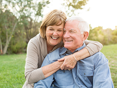 An elderly couple, a man and woman, embrace each other with smiles in a park setting.