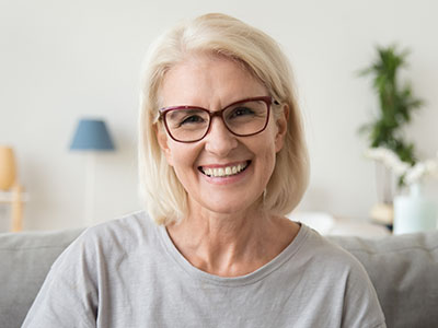 The image shows a smiling woman with short blonde hair, wearing glasses and a grey top, seated indoors on a couch.