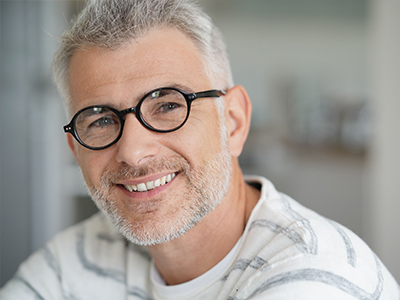 The image is a photograph of a middle-aged man with glasses, smiling at the camera. He has short hair and is wearing a white top with a patterned design.