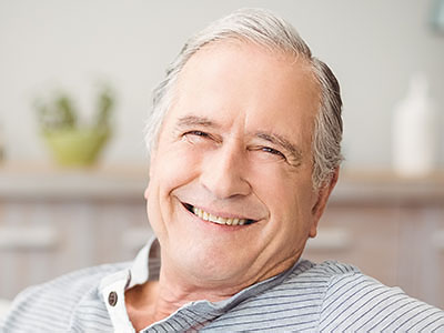 The image shows an elderly man with gray hair, wearing a blue shirt and glasses, smiling at the camera. He appears to be indoors, possibly in a living room or similar setting, as suggested by the presence of a white wall and what looks like a piece of furniture in the background.