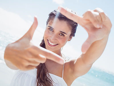 A young woman with long hair is smiling and holding up a finger in front of her face, creating a frame for the camera. She is outdoors during daylight, with a clear sky and beach setting visible behind her.